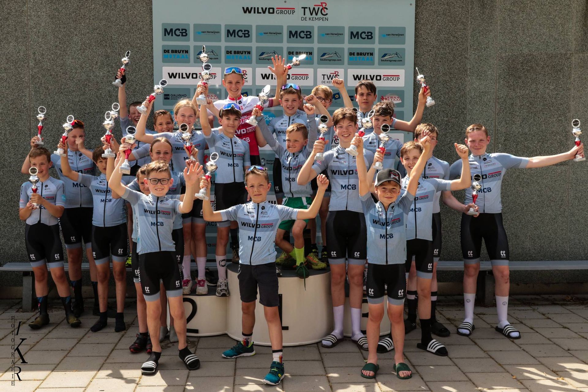 Een groep jonge wielrenners, gekleed in bijpassende shirts, viert hun overwinning op een podium en heft de trofeeën omhoog.