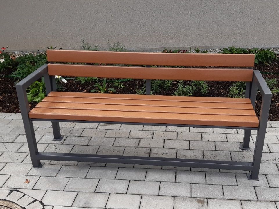 Eine Holzbank mit Metallrahmen steht auf einem gemauerten Gehweg