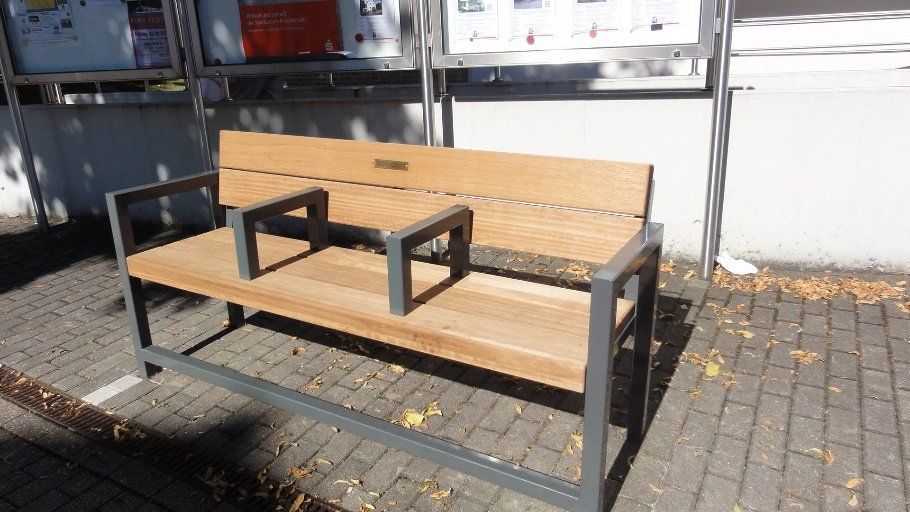 Auf einem gepflasterten Gehweg steht eine Holzbank mit Metallrahmen.