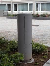 Ein grauer Zylinder steht mitten auf einer gepflasterten Auffahrt neben einem Gebäude.