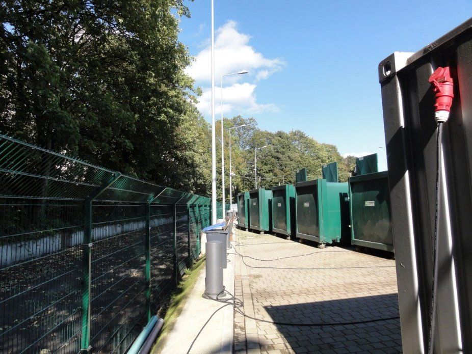 Eine Reihe von grünen Müllcontainern stehen hinter einem Zaun