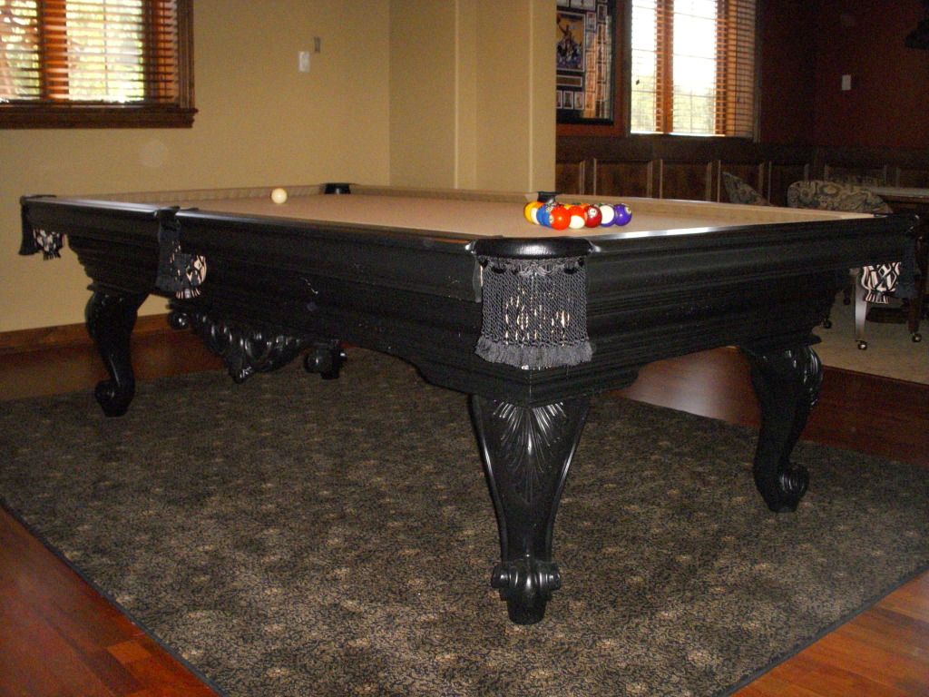 Black pool table with ornate legs, balls, and a rug.