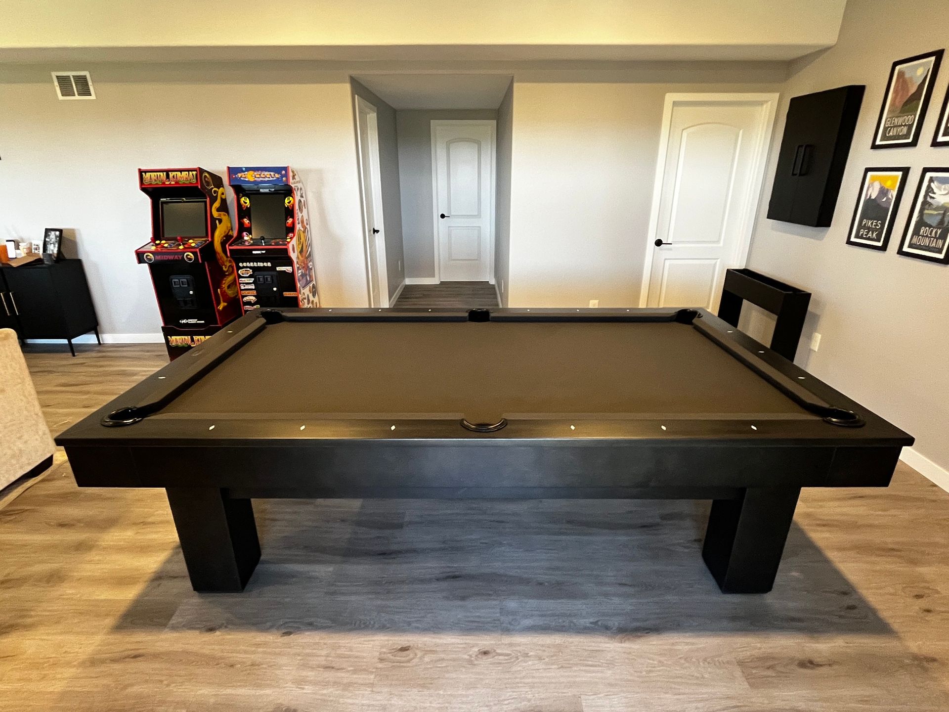 Black pool table in a finished basement, with arcade games, and framed artwork on the walls.
