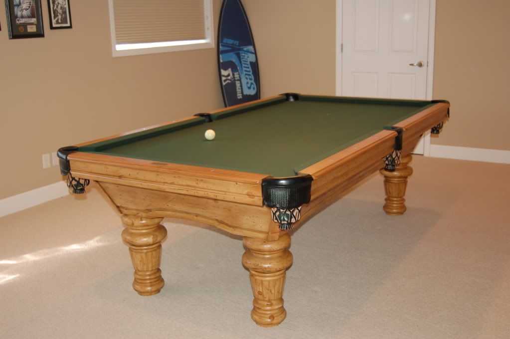Pool table with green felt, brown wood, and a white ball, in a room with a surfboard.
