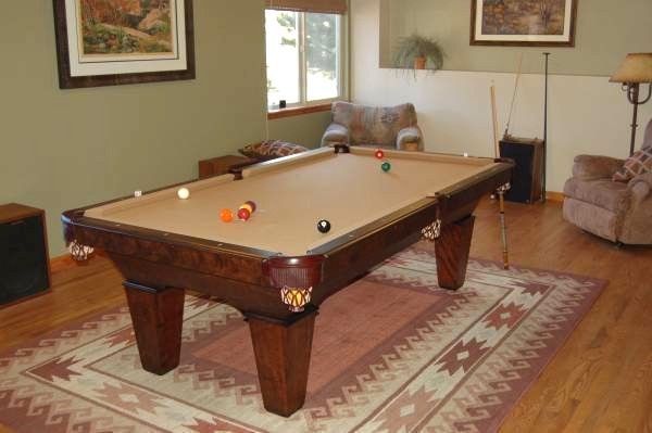 Pool table in a room, with balls scattered on tan felt, surrounded by a rug and chairs.