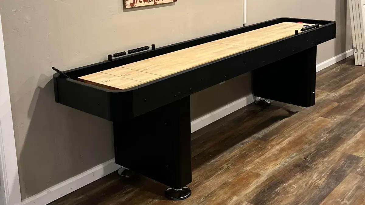 A black shuffleboard table with a light wood playing surface, positioned against a wall. The table sits on wheels.