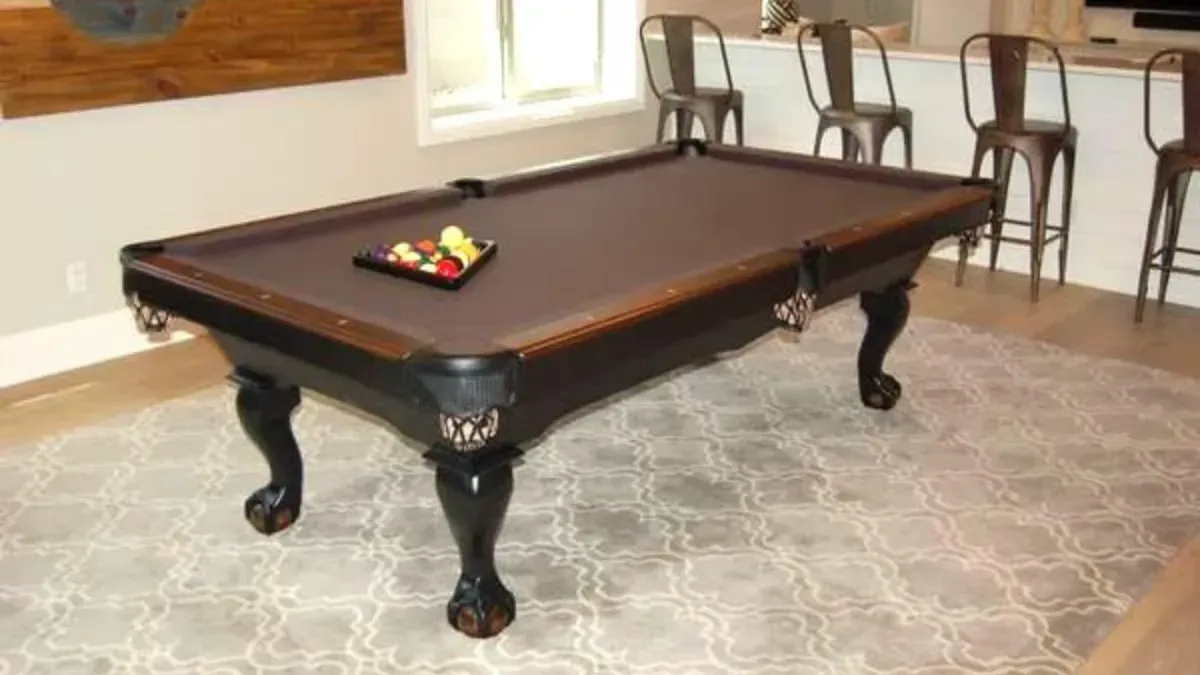 Brown pool table in a room, balls racked, with bar stools in the background.