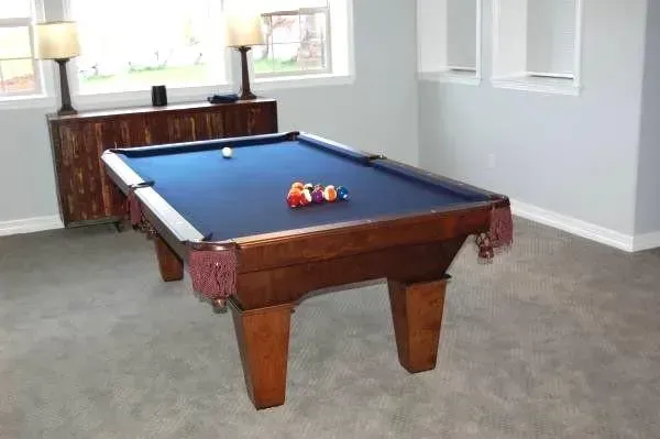 Pool table with blue felt, wooden frame, and balls, in a room with a window, lamps, and a carpet.