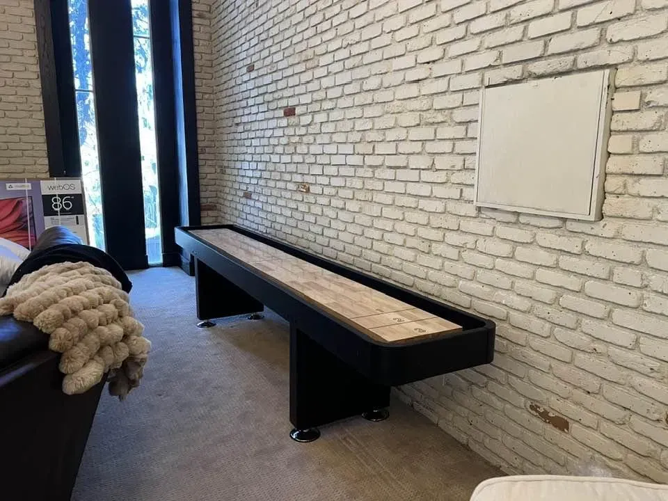 Shuffleboard table in a room with a brick wall. The table is black with a wood surface.