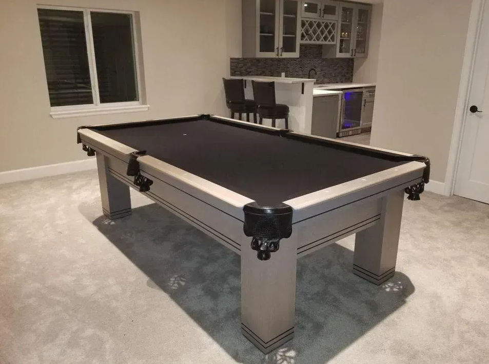 Gray pool table with black felt in a finished basement with a bar.
