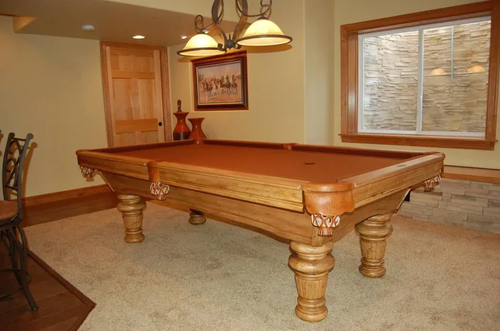 A wooden pool table in a finished basement with beige walls, a window, and overhead lighting.