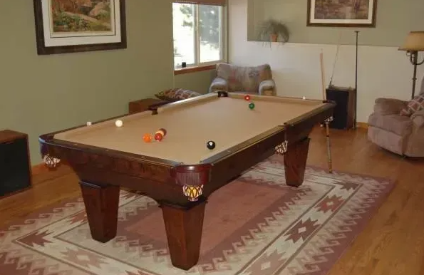 Pool table in a room; balls scattered on the felt. Brown table, tan felt, green walls, rug, and wood floor.