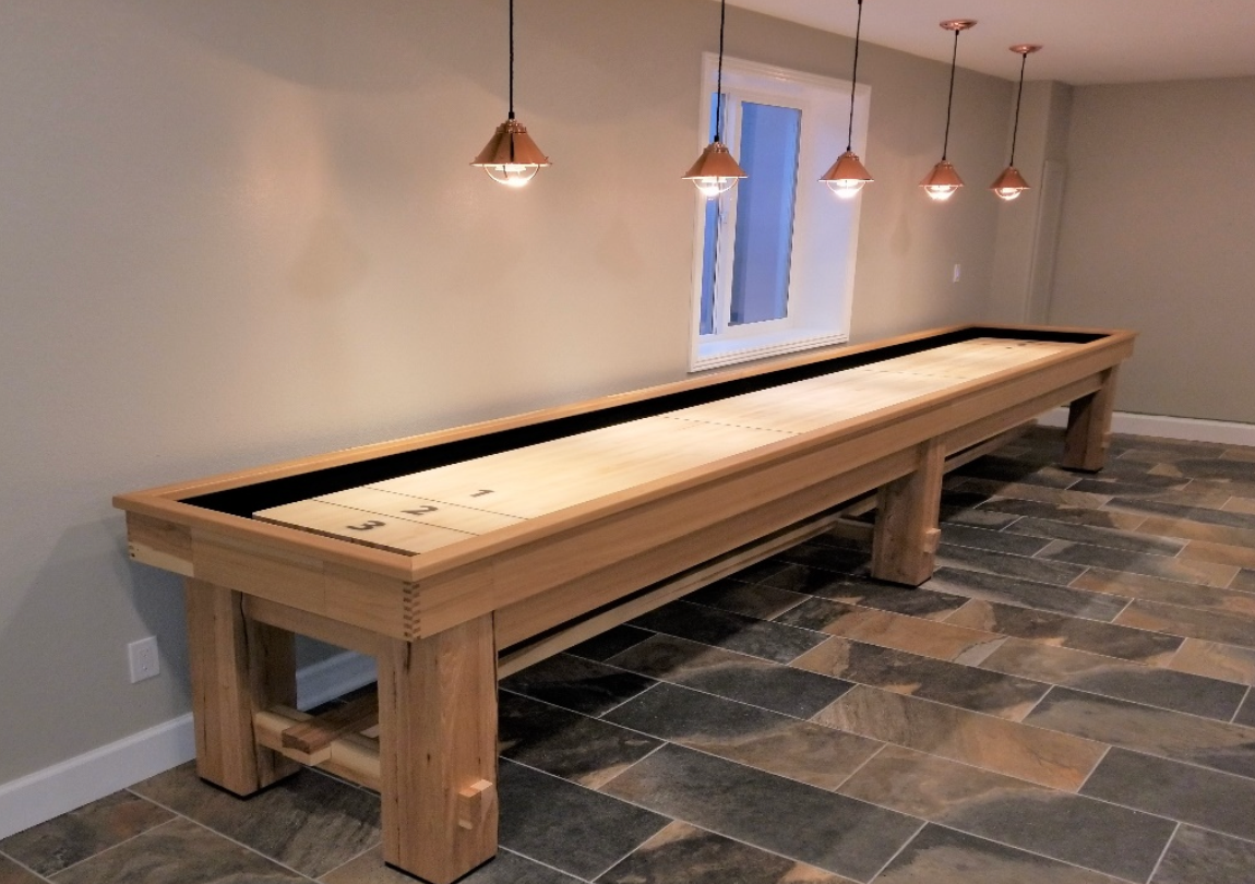 A long wooden shuffleboard table in a room with a window and pendant lights.