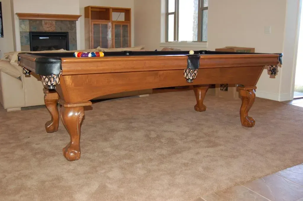 Wooden pool table with curved legs, balls arranged, in a room with a fireplace and carpet.