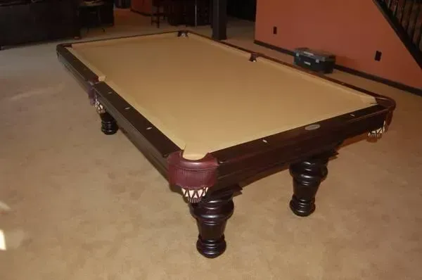 A brown pool table with tan felt, four dark wood legs, on a beige carpet.