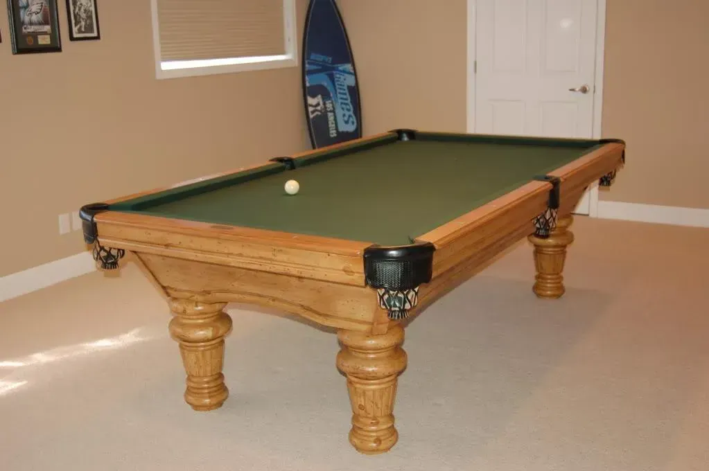 Pool table with green felt, light wood, and a single white ball, in a room with light walls and a surfboard.