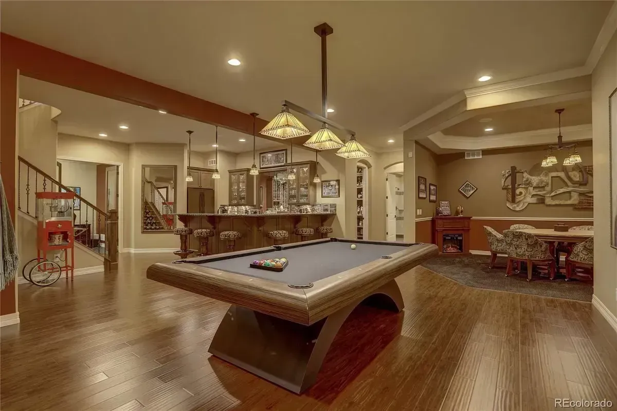 Pool table in a game room with a bar, fireplace, and dining area. Wooden floors and warm lighting.
