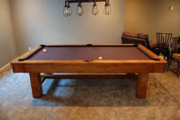 Pool table with brown felt and wooden frame, centered in a room with a light fixture above.