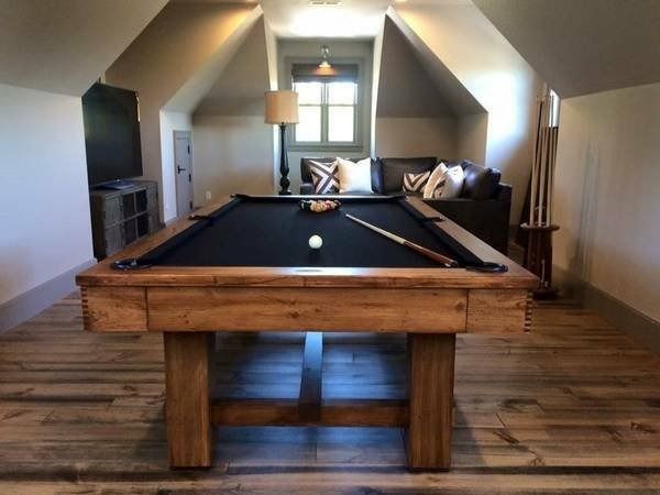 Wooden pool table in an attic room with couch, TV, and window.