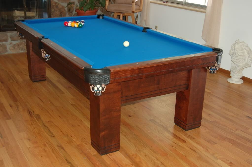 Wooden pool table with blue felt, balls arranged, white cue ball, set in a wood-floored room.