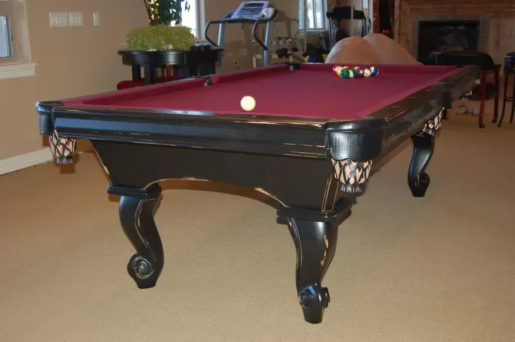 A black pool table with red felt, a white cue ball, and assorted colored balls sits in a room with tan carpet.