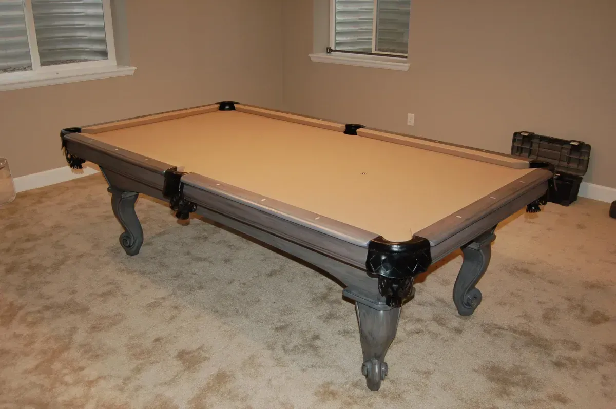 Pool table in a beige room with light-colored carpet and small windows. The table is gray with a tan playing surface.