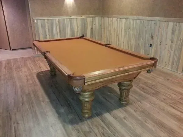 Pool table with brown felt on wooden floor, surrounded by wood paneled walls.
