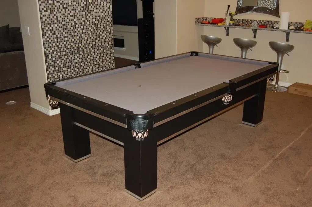 Black pool table with gray felt in a room. Three bar stools and a patterned wall.