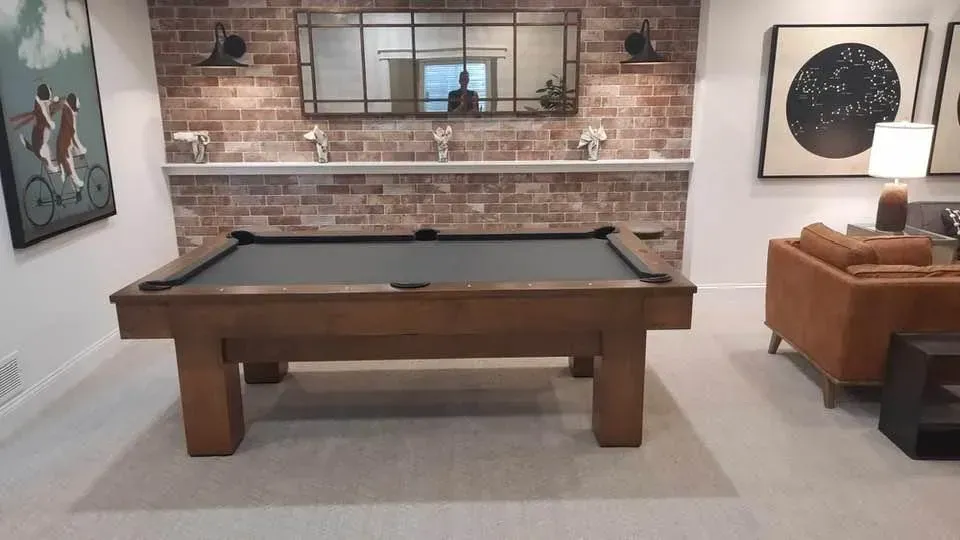 Pool table in a room with a brick wall, mirror, and artwork. A person is visible in the mirror.