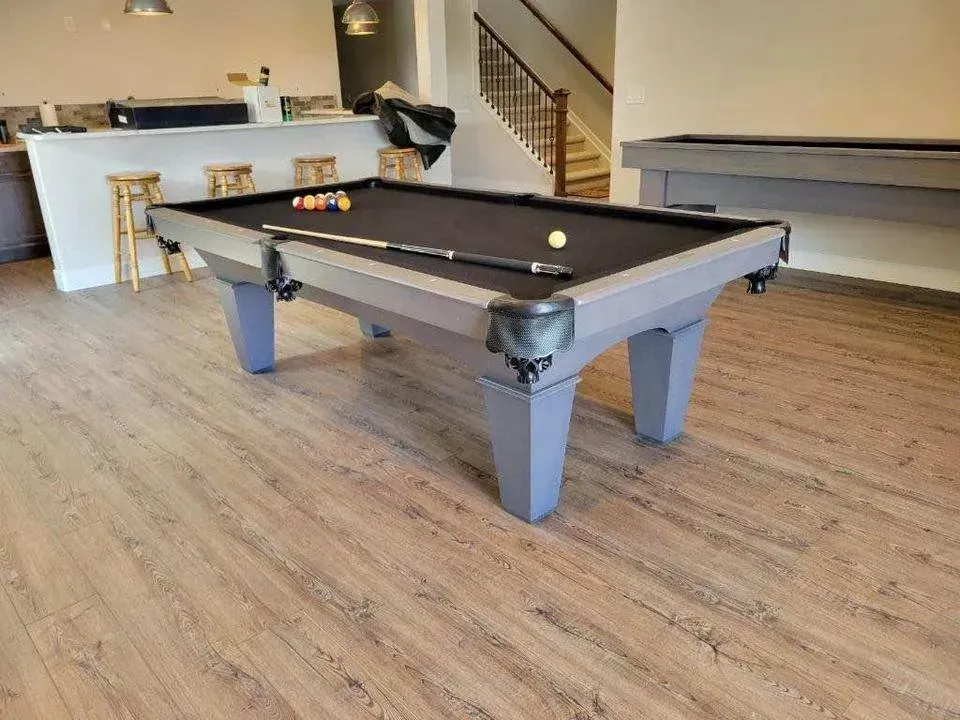 Pool table in a room, with balls, cue stick, and a bar area in the background. Wooden floors.