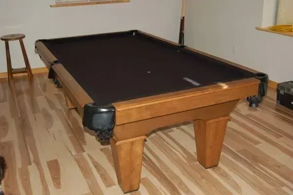 A brown pool table in a room with hardwood flooring, with a stool nearby.
