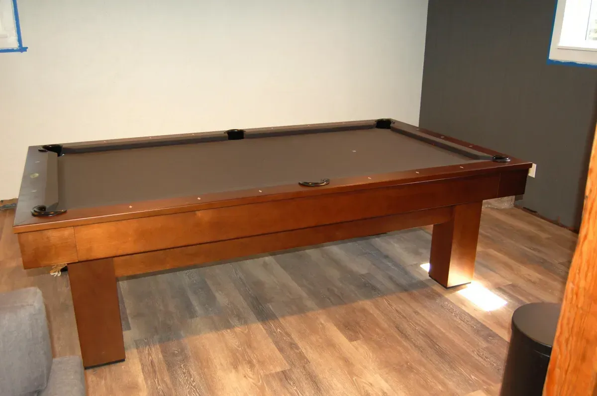 A brown pool table in a room with wood flooring, and gray and white walls.