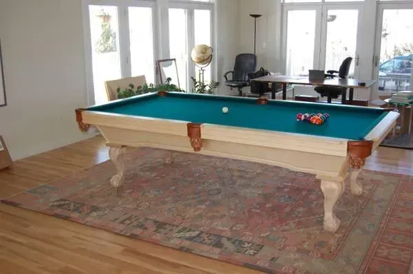 Pool table with green felt, white frame, and balls on a patterned rug in a bright room.