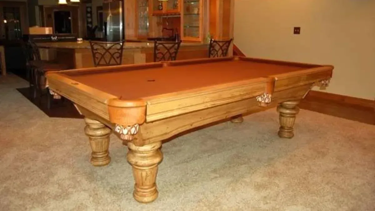 Wooden pool table with brown felt in a room with a bar in the background.