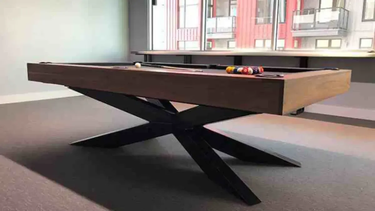 Pool table with brown wood top and black cross-shaped legs, balls on the felt, in front of a window.