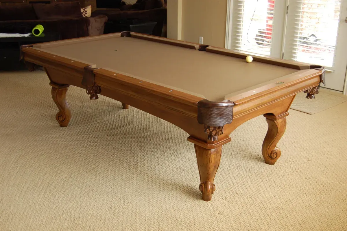 Pool table with a brown felt top, ornate wooden frame and legs, and a white ball on the table.
