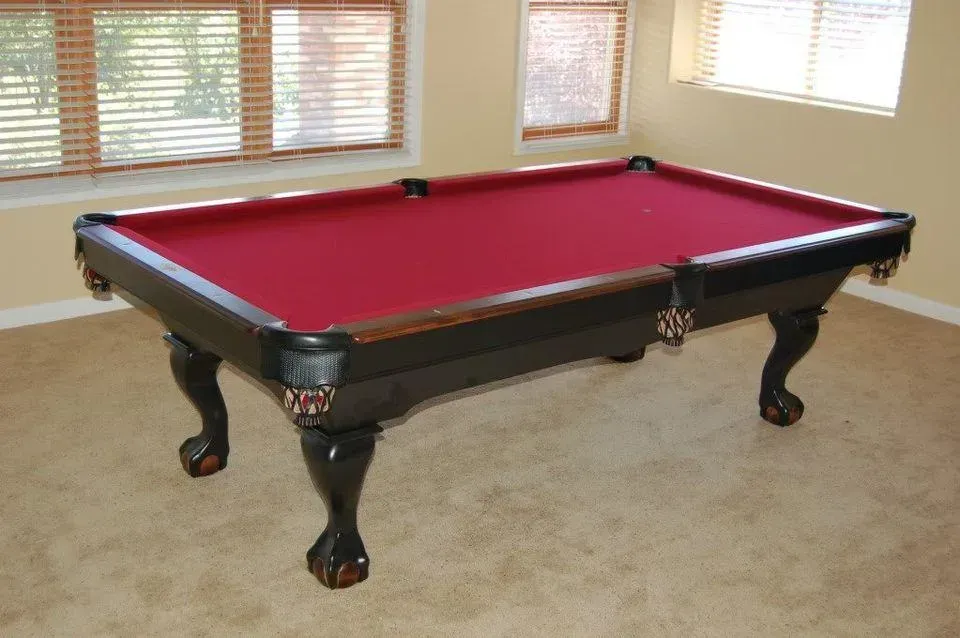 Pool table with red felt top, black frame, and ornate legs in a beige room with windows.