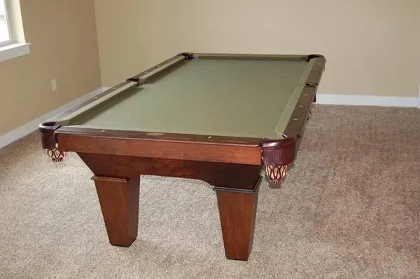 Pool table in a room with tan walls and carpet. The table is brown with a green felt surface.