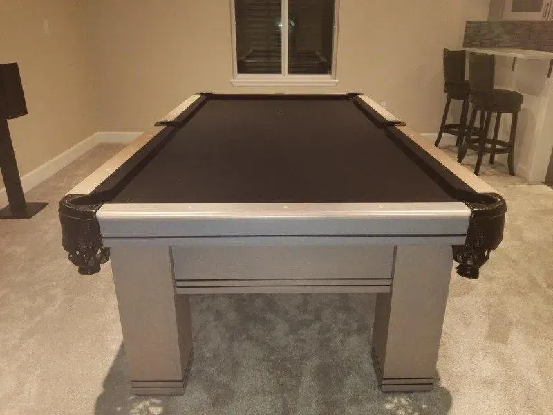 Pool table with black felt, gray frame, in a well-lit room, with stools and a window in the background.