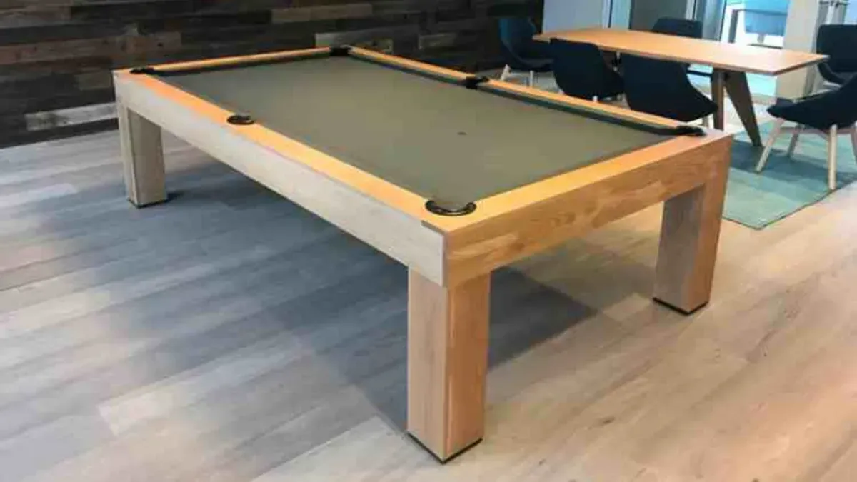 Pool table with light wood frame, green felt, and black pockets. The table sits on a light gray wooden floor.