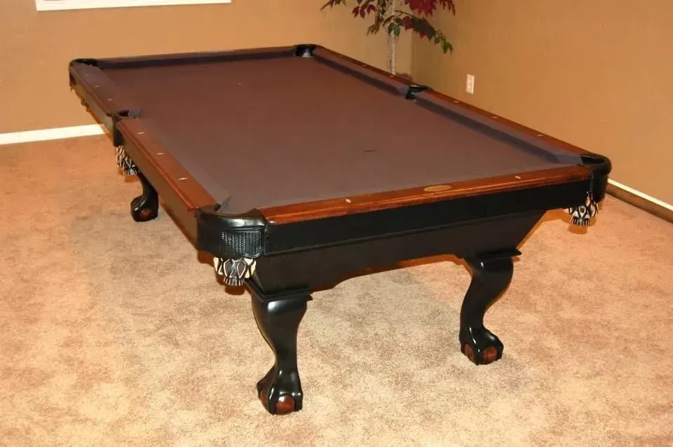 Pool table in a room with brown carpet and beige walls. Black legs, brown trim, and gray felt top.