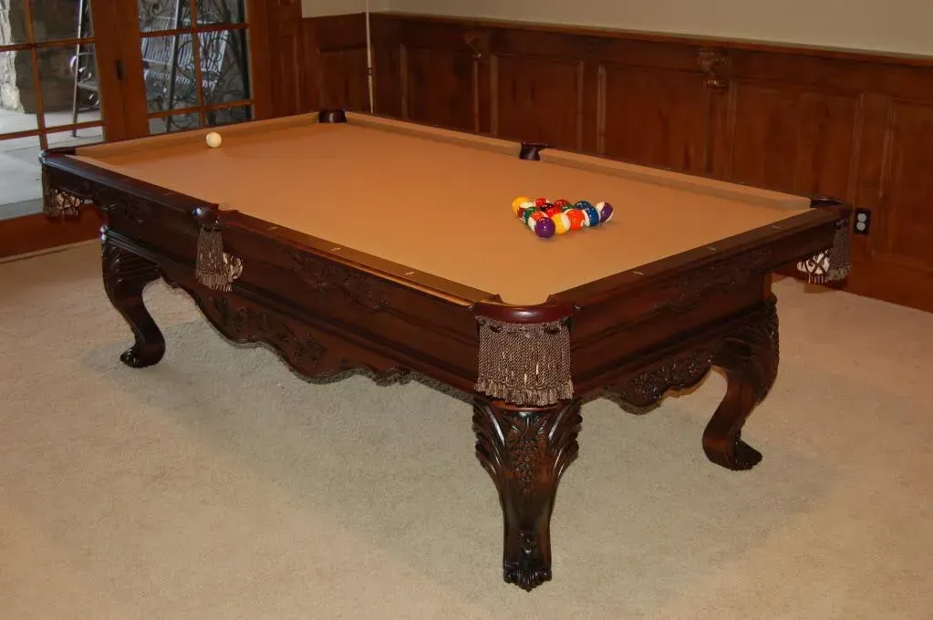 A pool table with a tan felt top and carved dark wood frame, balls arranged, in a room.