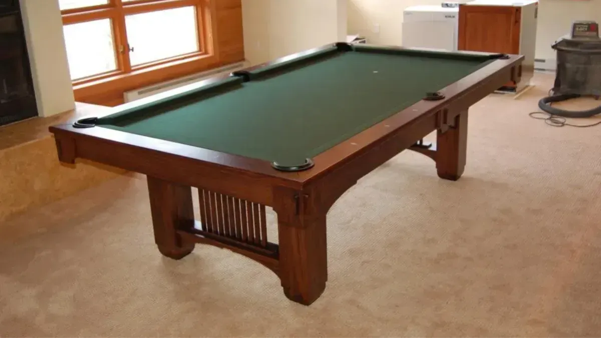 A wooden pool table with green felt sits in a room with a window and a neutral carpet.