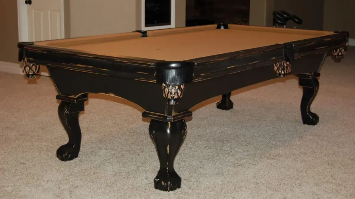 Black pool table with ornate legs, tan felt, set in a room with tan carpet.