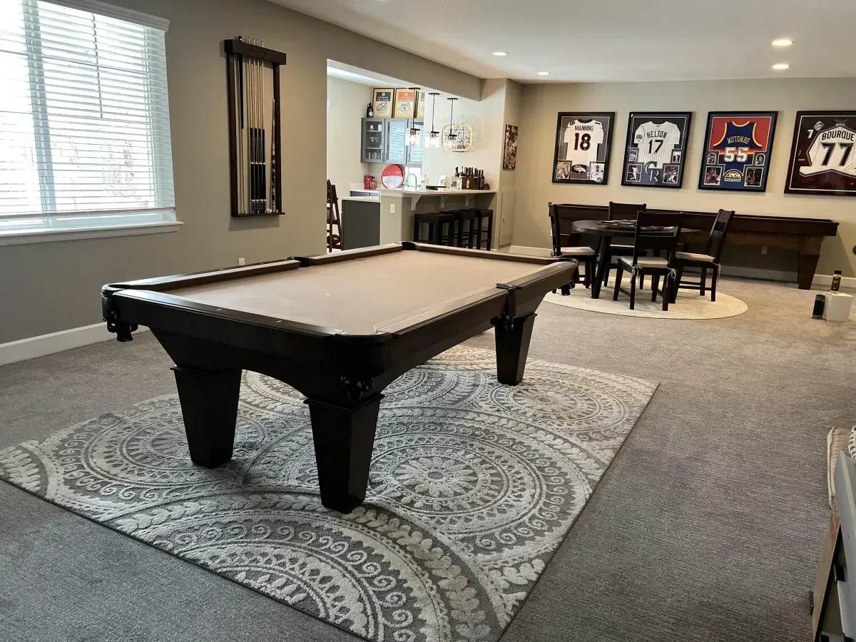 Pool table on patterned rug in a basement game room with seating, framed jerseys, and a bar area.