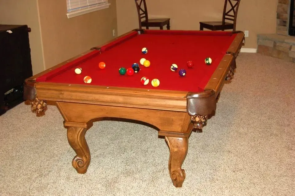 Pool table with red felt, scattered balls, and ornate wood legs in a room with tan walls and carpet.