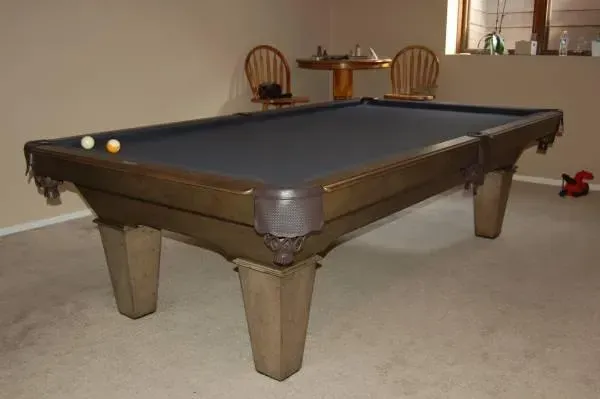 A brown pool table with dark blue felt, two balls, and two wooden chairs in a room.