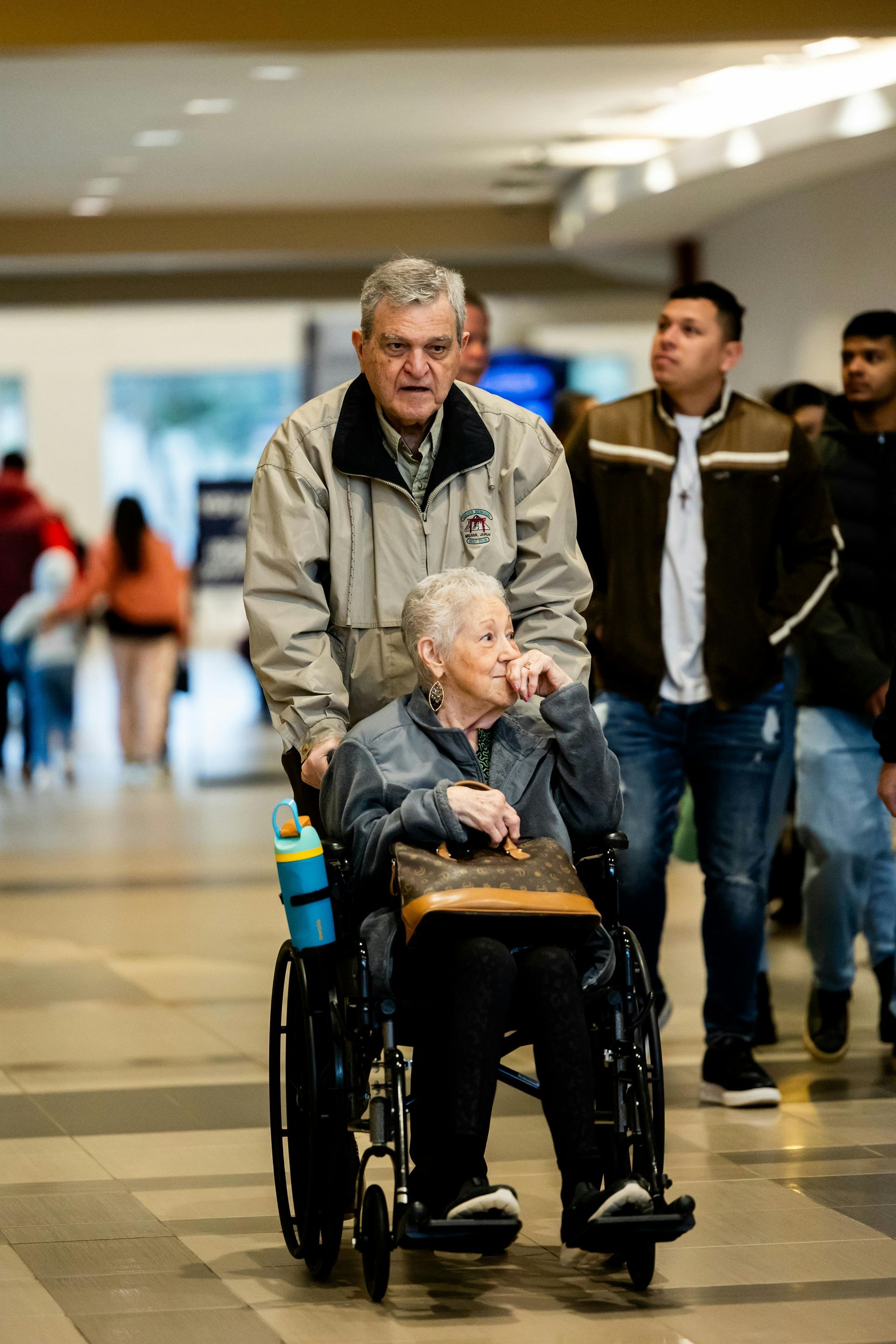 Man pushing a person in a wheelchair through a public space; other people are walking nearby.