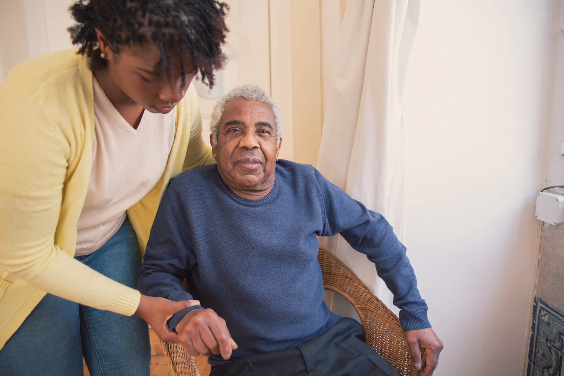 Caregiver assisting an older adult in a blue sweater to sit in a chair, offering comfort and companionship. Interior setting, natural light.