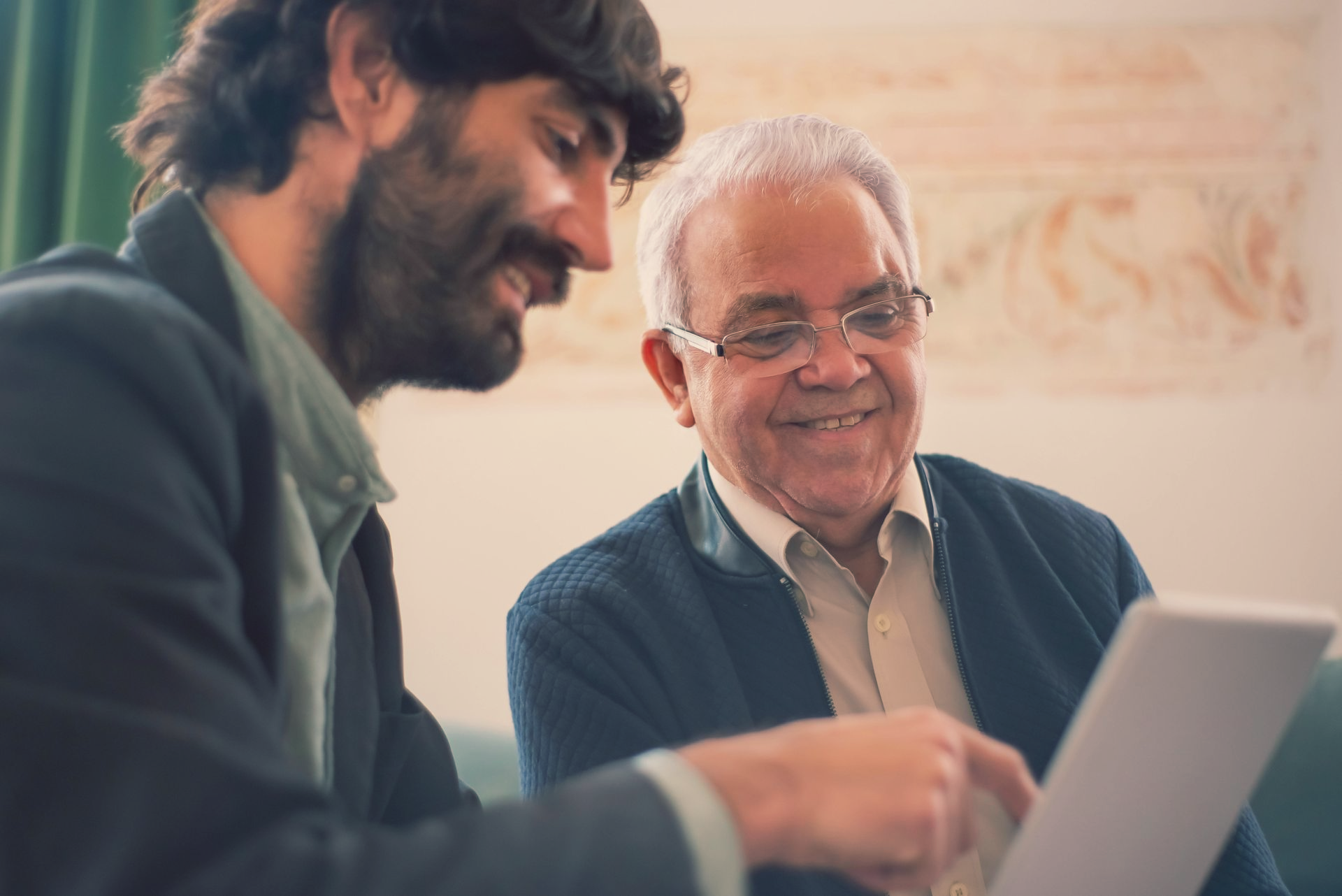 Man points at tablet screen, smiling with an older man, in a room.
